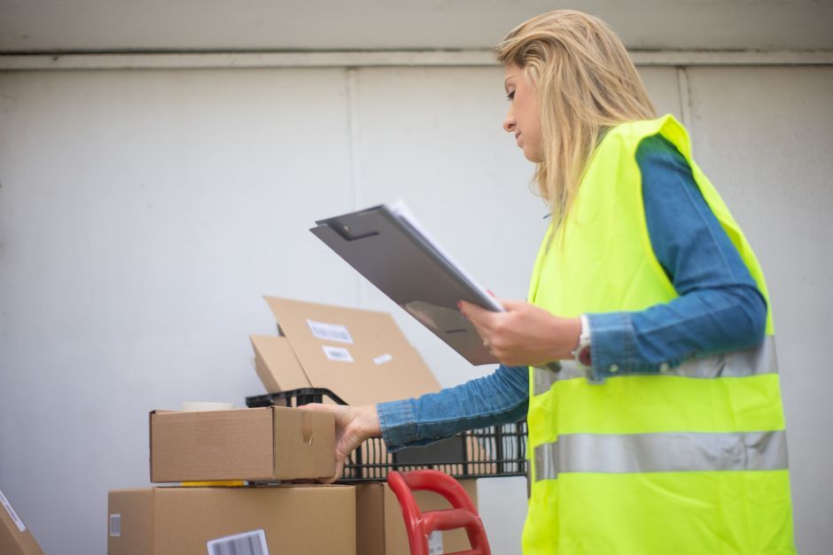 Woman in high-visibility vest handling boxes for delivery service inside a warehouse setting.